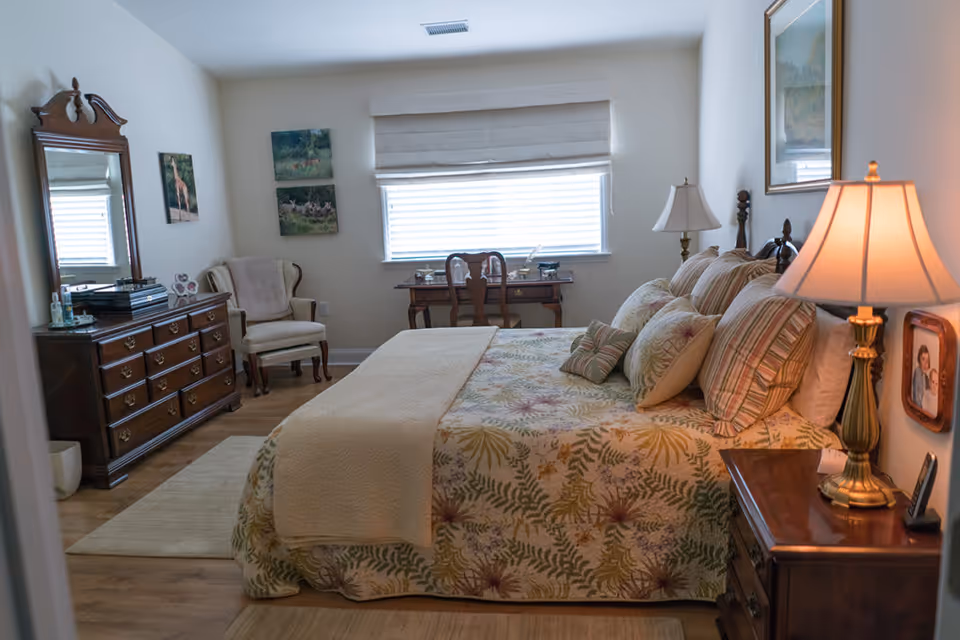 A cozy bedroom with a large bed covered in a floral patterned bedspread and multiple pillows. There is a wooden nightstand with a lamp on the right side of the bed. Across from the bed is a wooden dresser with a mirror and various items on top. A cushioned armchair is placed near the dresser. A wooden desk with a chair is positioned under a window with blinds, and two paintings hang on the wall above the armchair. The room has wooden flooring and light-colored walls.