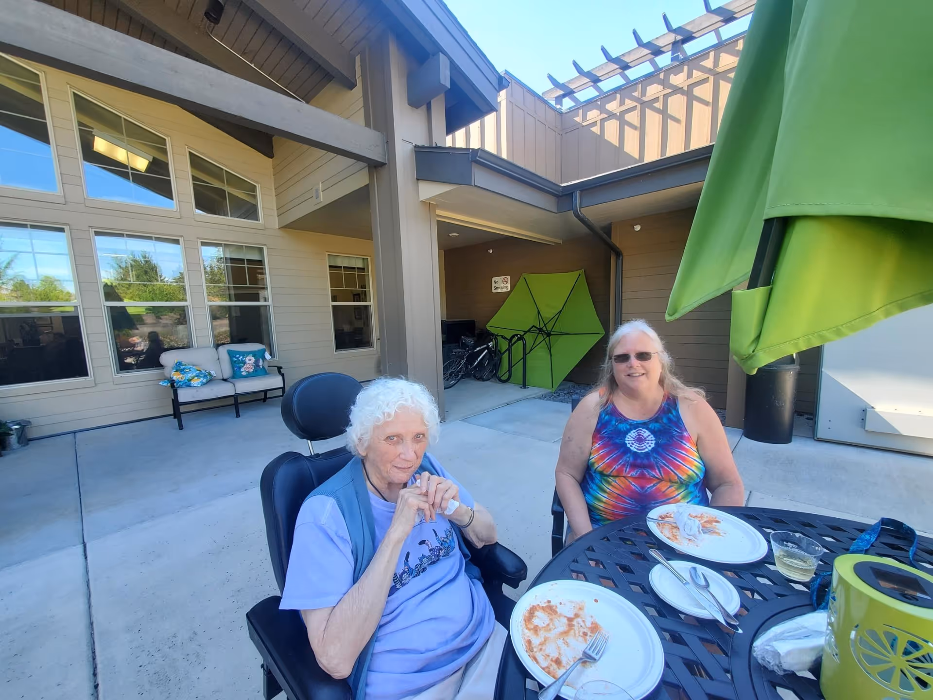 Two elderly women sitting at a round outdoor metal table with empty plates and utensils, enjoying a sunny day at an outdoor patio area of a senior living facility. One woman has white curly hair and is seated in a wheelchair, wearing a light purple shirt and blue vest. The other woman has long blonde hair, glasses, and is wearing a colorful tie-dye tank top. Behind them is the building exterior with large windows, a bench with cushions, and a green umbrella.
