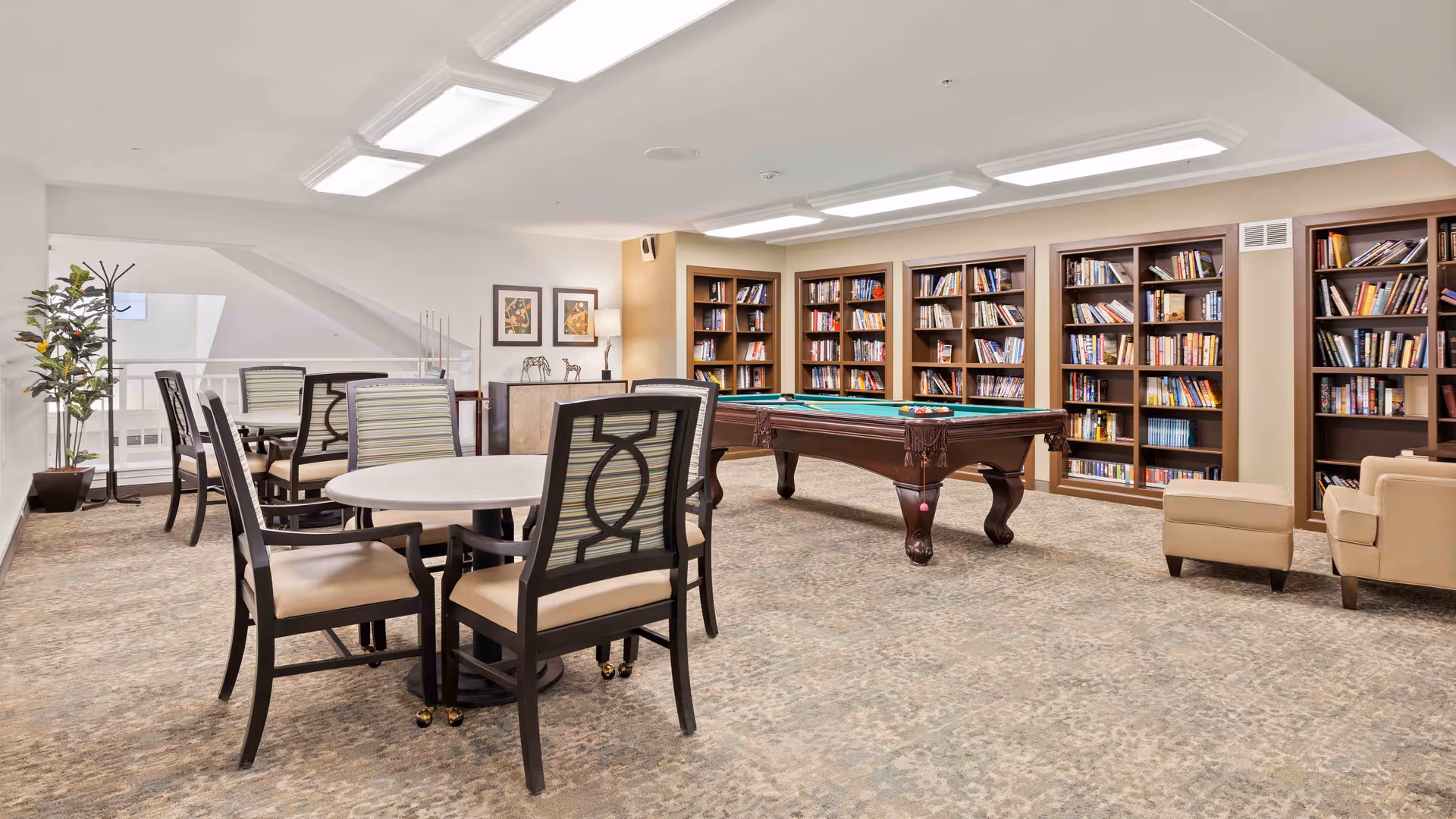 A spacious room with a pool table in the center, surrounded by bookshelves filled with books. There are several round tables with chairs arranged around them, and a couple of armchairs with an ottoman near the bookshelves. The room has bright overhead lighting and a carpeted floor, creating a comfortable and inviting atmosphere.