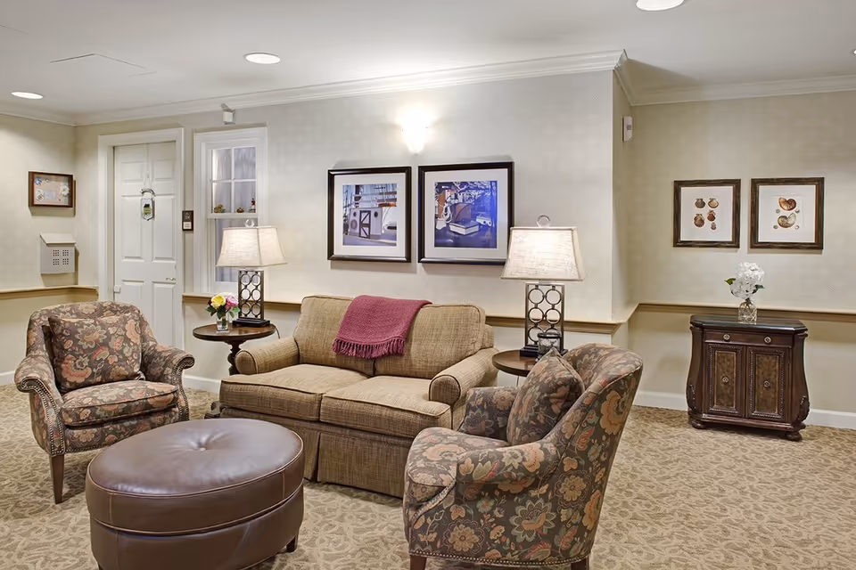 Cozy seating area in a senior living common room with a sofa, patterned armchairs, ottoman, side tables with lamps, framed artwork, and a small cabinet.
