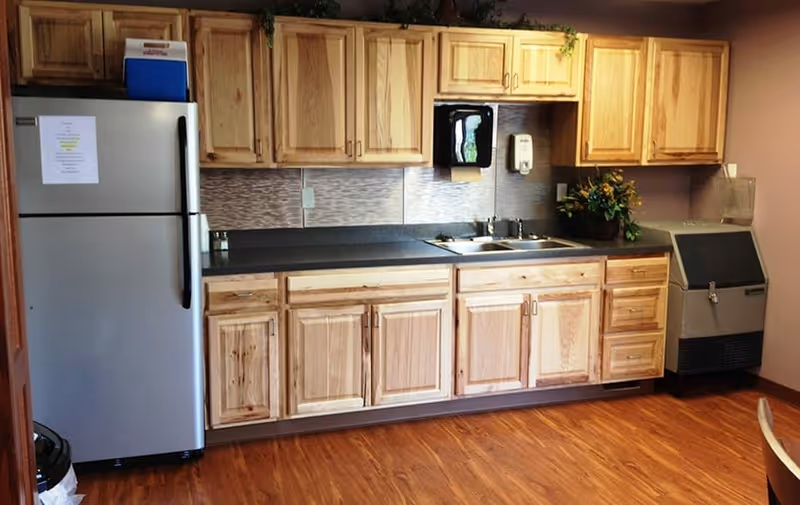 A kitchen area with wooden cabinets, a stainless steel refrigerator, a double sink, a black countertop, a wall-mounted soap dispenser, and an ice machine. The floor is wood laminate, and there is a small plant decoration on the counter.