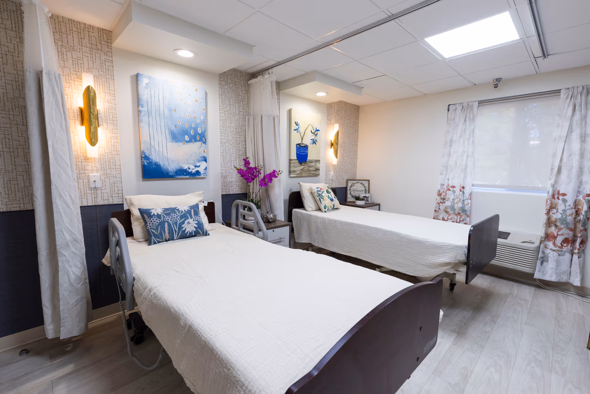 A bright shared patient bedroom with two adjustable beds, bedside tables, decorative artwork and a window with floral curtains.