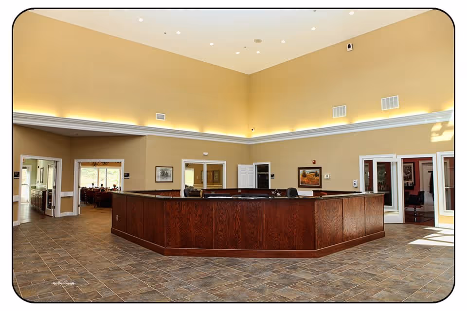 Spacious interior area of a senior living community featuring a large, dark wooden reception desk in the center. The room has high ceilings with recessed lighting and beige walls. Multiple doorways lead to other rooms, including a dining area visible through one doorway. The floor is covered with brown and gray tile.
