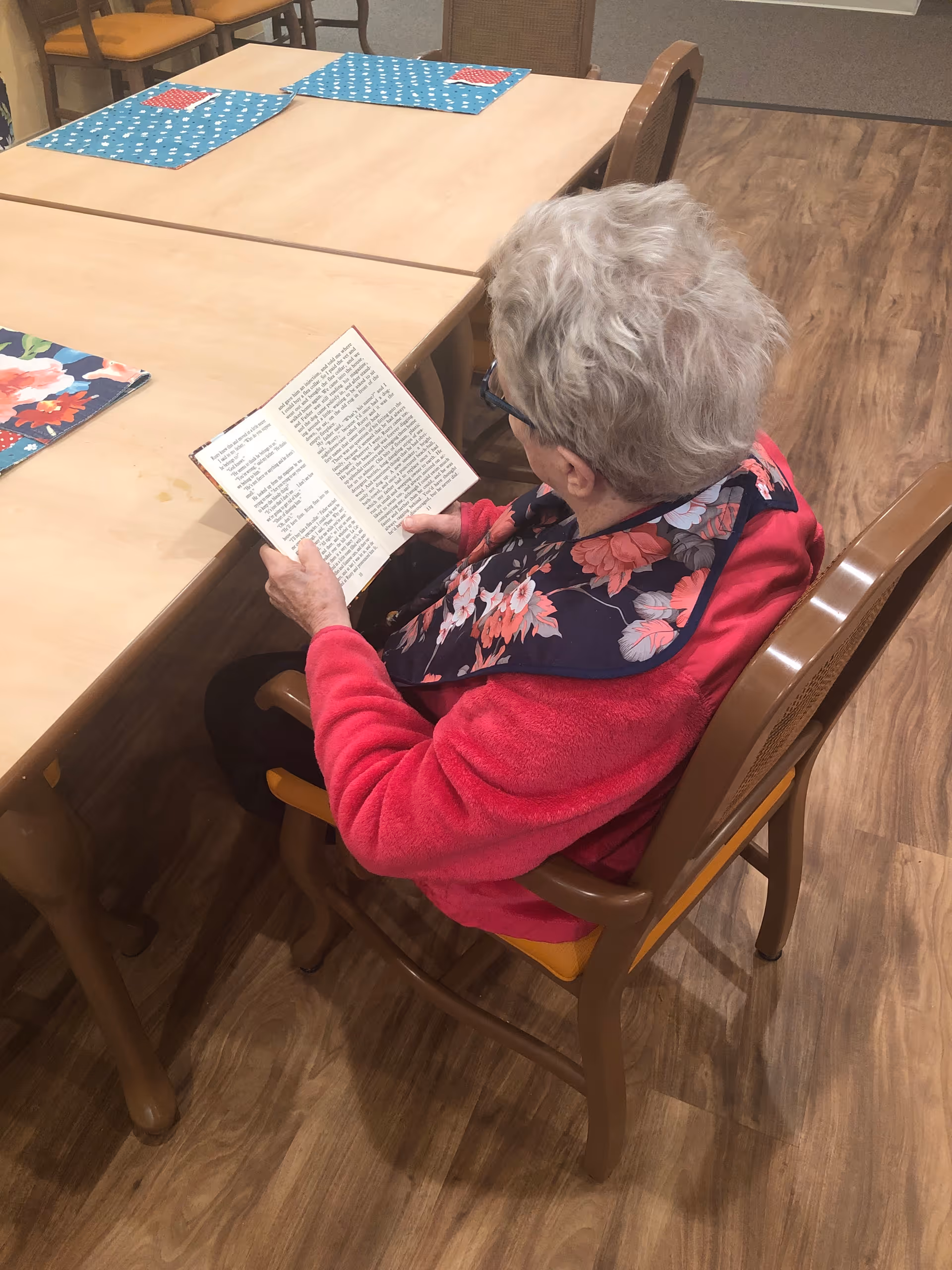 A senior woman in a pink jacket sits at a dining table reading a book.