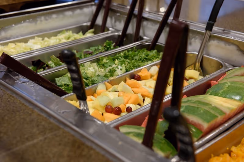 A salad bar with various fresh ingredients including chopped lettuce, leafy greens, mixed fruit pieces with grapes, and sliced watermelon, each in separate metal containers with serving utensils.