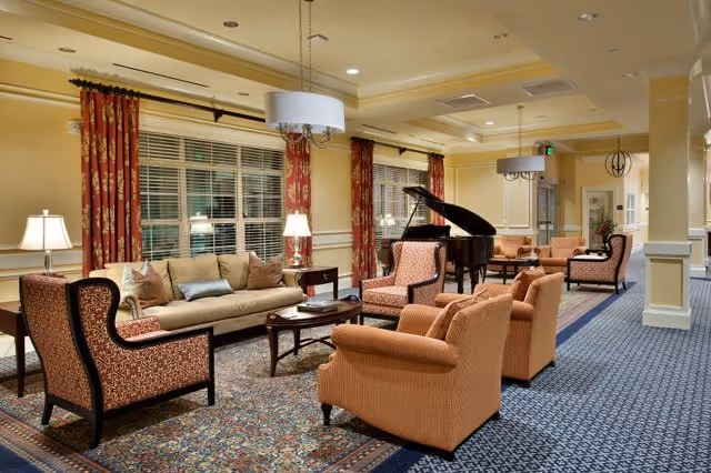Spacious furnished lounge with sofas, armchairs, coffee tables, patterned rugs, and a grand piano under warm lighting.