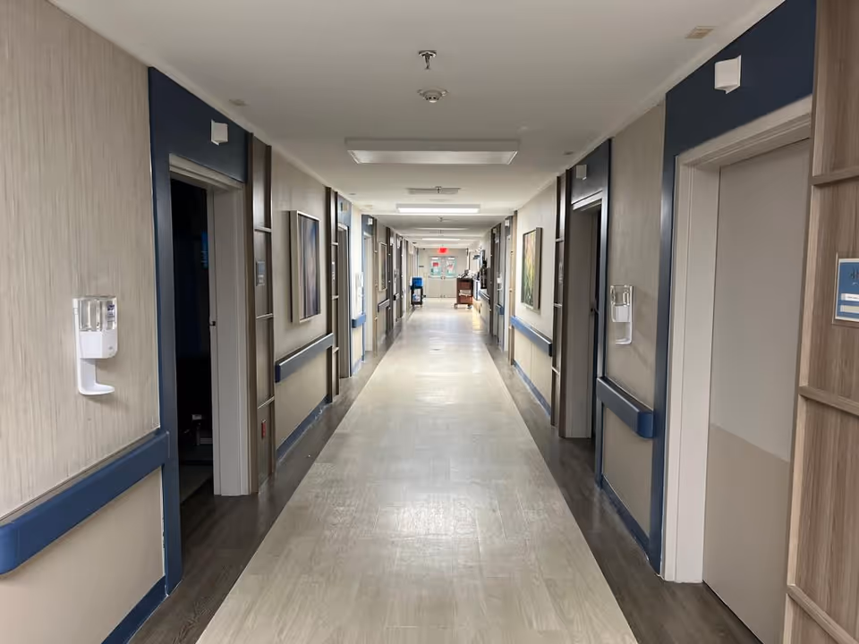 Long, well-lit hallway in a care facility with multiple patient room doors, handrails, and wall-mounted dispensers.
