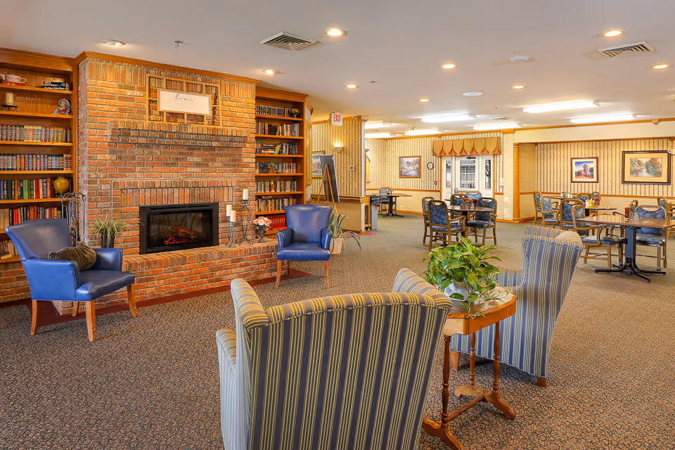 Spacious common room with a brick fireplace, bookshelves, upholstered armchairs, and dining tables.