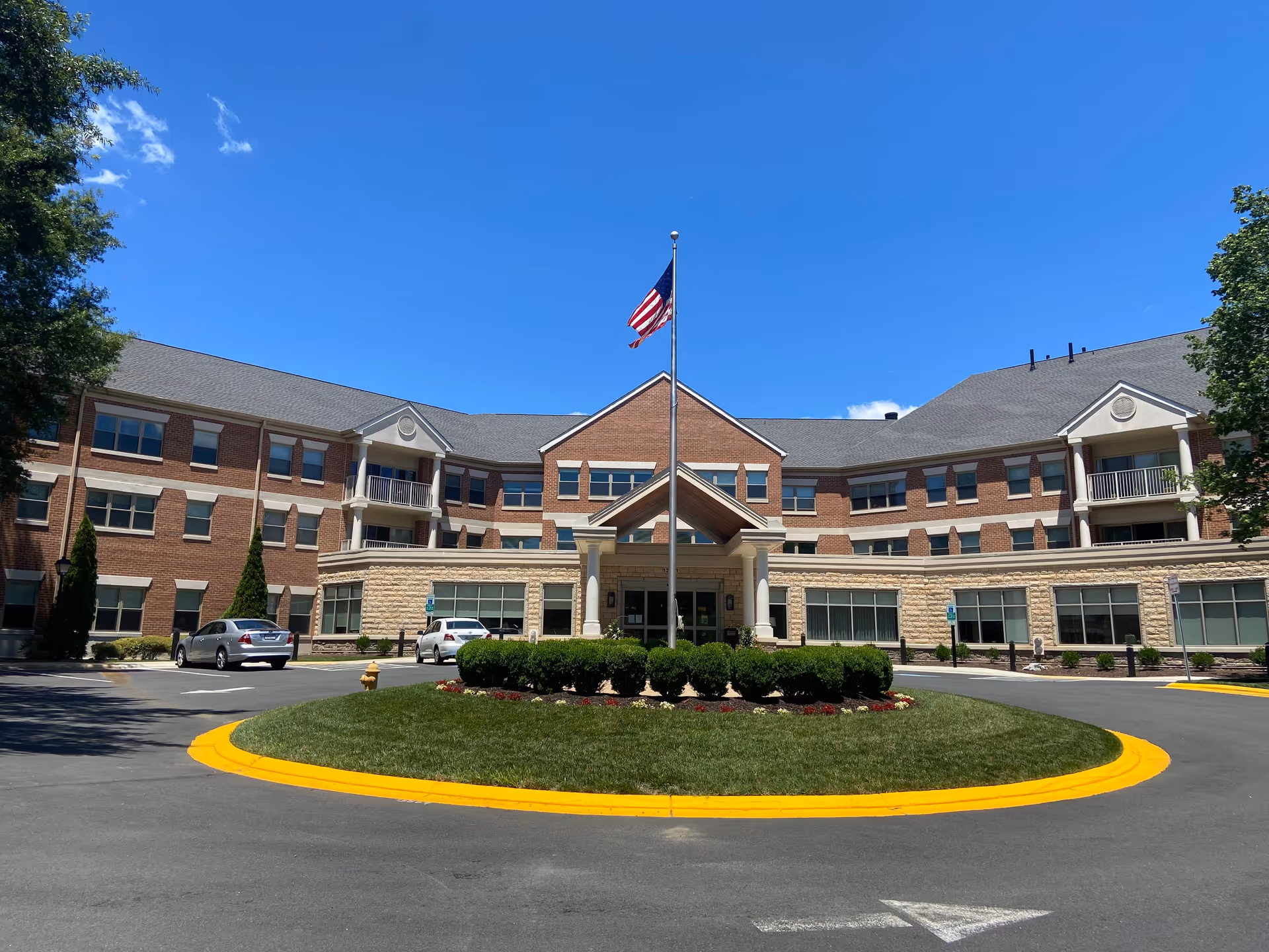 Front exterior view of Westminster at Lake Ridge a CCRC building with a circular driveway, landscaped greenery, and an American flag on a flagpole in the center under a clear blue sky.