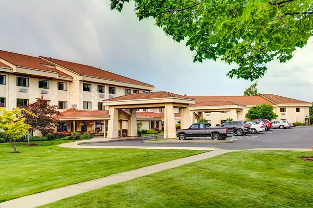Exterior view of Grande Ronde Retirement Residence showing a multi-story building with a covered entrance, a parking lot with several cars, and well-maintained green lawns and trees in the foreground.