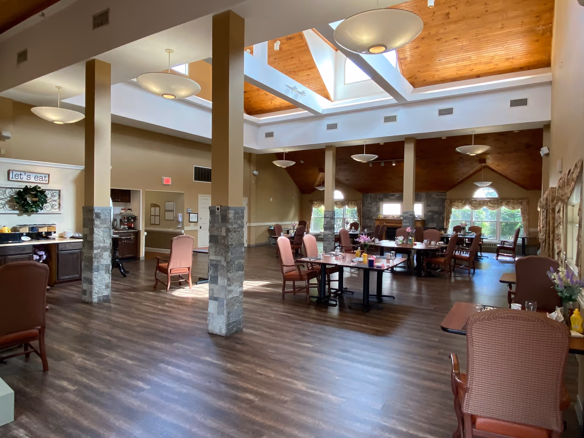Spacious dining room with wooden floors, multiple tables and chairs arranged for meals, large windows letting in natural light, and a high ceiling with wooden paneling and modern light fixtures. A sign on the wall reads 'let's eat'.