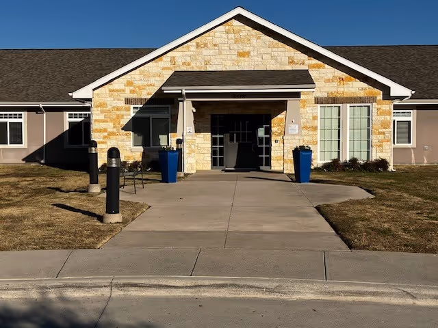 Front exterior view of a single-story building with a stone facade and a peaked roof. There is a concrete walkway leading to the entrance, flanked by two large blue planters. The building has multiple windows and a clear blue sky above.