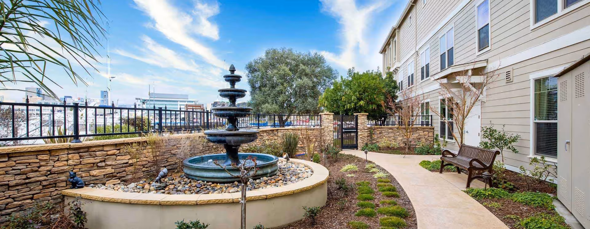 Outdoor courtyard area at Oakmont of Mariner Point featuring a multi-tiered water fountain surrounded by a stone wall and landscaping. There is a paved walkway with green plants and a wooden bench along the side of a beige building under a blue sky with some clouds.