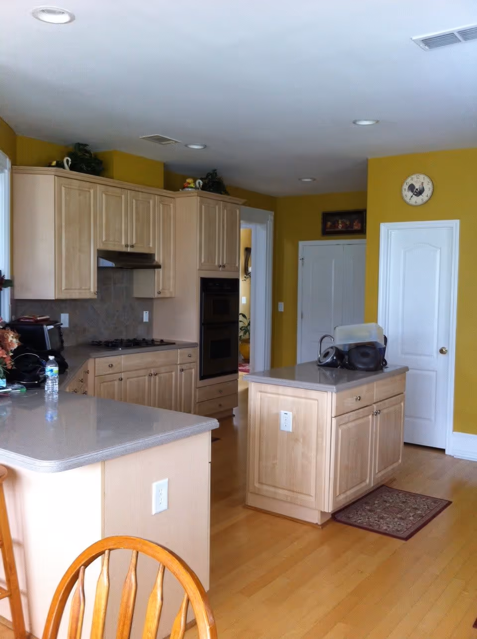 A kitchen with light wood cabinets and a central island. The walls are painted yellow, and the floor is light wood. There is a stove with a range hood, a double oven, and various kitchen appliances on the counters. A clock with a rooster design is mounted on the wall above a white door. A wooden chair is partially visible in the foreground.