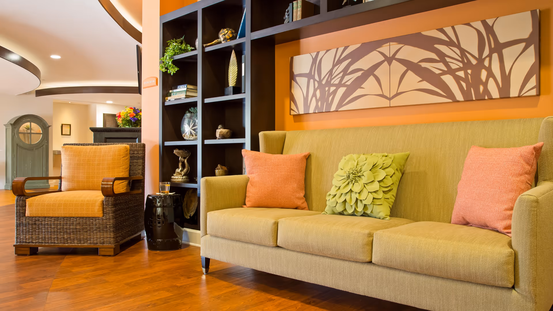 A cozy living room area with a beige sofa adorned with two coral-colored pillows and one green floral pillow. Next to the sofa is a wicker armchair with an orange cushion. Behind the sofa is a decorative wall art piece with a nature-inspired design. A black shelving unit with various decorative items is visible, along with a small black side table holding a glass of water. The room has warm wooden flooring and soft lighting.