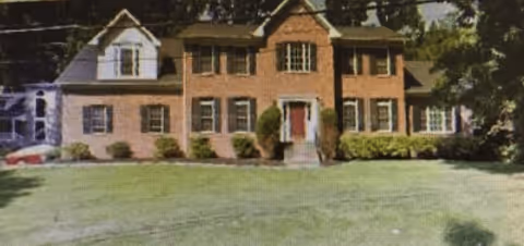 A large two-story brick house with multiple windows and a central front door, surrounded by a well-maintained lawn and some shrubs. Trees are visible in the background.