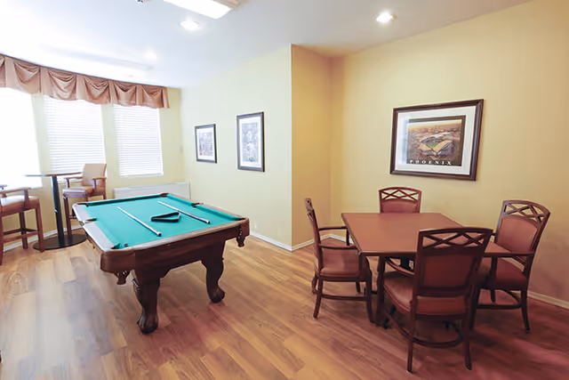 A well-lit room with a pool table in the center, a small round table with four chairs to the right, and two high chairs near the window with blinds and a valance. The walls are painted light yellow and decorated with framed pictures.