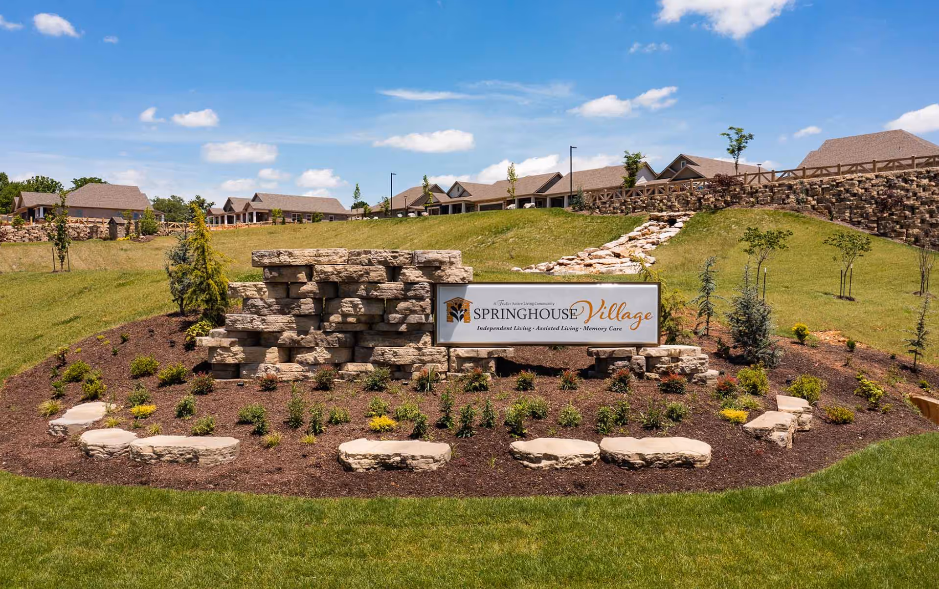 Outdoor landscaped area with a stone sign that reads 'Springhouse Village Independent Living • Assisted Living • Memory Care' under a blue sky with scattered clouds. Residential buildings are visible in the background on a grassy hill.