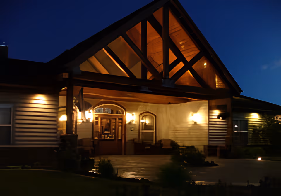 Exterior nighttime view of the entrance to a building with warm lighting illuminating the porch area and wooden architectural details under a peaked roof.