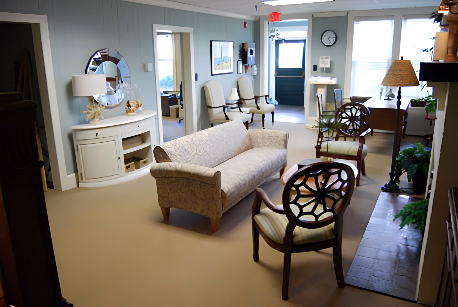 A cozy living room area in a senior living facility with a beige patterned sofa, two wooden chairs with decorative backs, a white sideboard with a round mirror above it, a floor lamp, and a desk near large windows letting in natural light. The walls are painted light blue, and there are several plants and framed pictures on the walls.