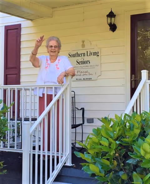 A smiling elderly woman waves from the front porch of a house beside a 'Southern Living For Seniors' sign.
