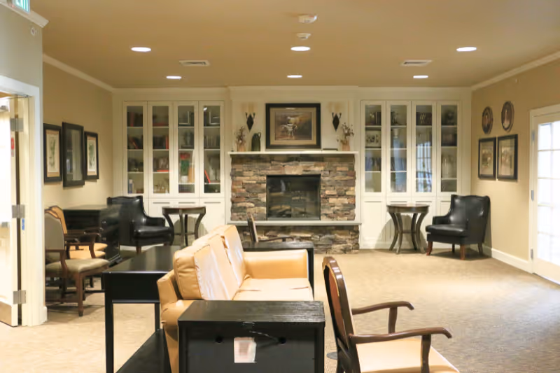 A cozy living room area with beige walls and carpet, featuring a stone fireplace in the center flanked by white built-in cabinets with glass doors. The room has various seating options including tan leather sofas, black armchairs, and wooden chairs. There are framed pictures on the walls and small tables next to the chairs. A door with glass panes is visible on the right side.