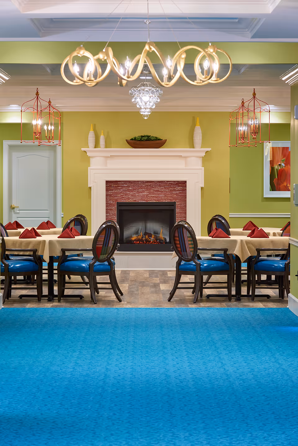 A dining room with tables covered in beige tablecloths and set with red folded napkins. The chairs have blue seats and striped backs. A fireplace with a red brick interior and white mantel is centered on the far wall, flanked by decorative vases and a bowl with greenery. The walls are painted green, and there are multiple light fixtures including a modern chandelier and two red cage-style pendant lights. A blue carpet covers the floor in the foreground.
