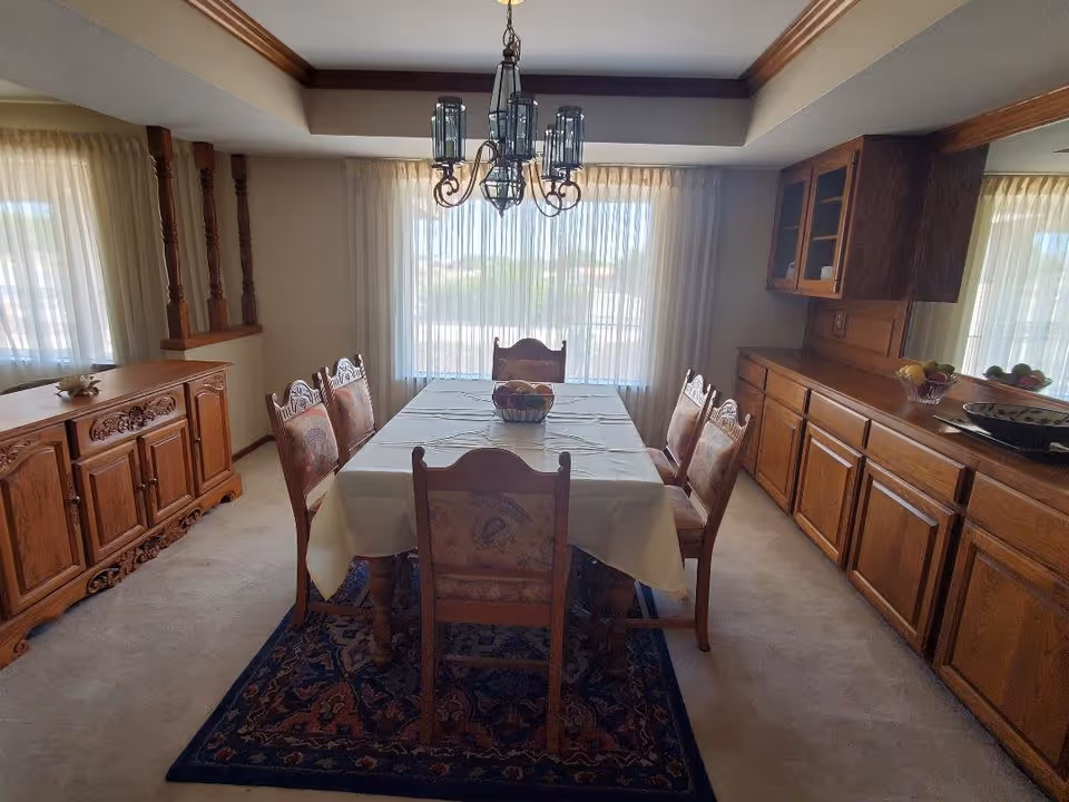 A dining room with a rectangular table covered with a white tablecloth and six wooden chairs with patterned upholstery. The room features large windows with sheer curtains, wooden cabinetry along one wall, a wooden sideboard, and a decorative chandelier hanging above the table. A patterned rug is underneath the table and chairs.