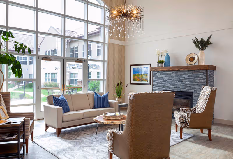 A bright and spacious living room area with large floor-to-ceiling windows showing an outdoor view of a building and greenery. The room features a beige sofa with blue pillows, two patterned armchairs, a wooden coffee table, a modern chandelier, and a stone fireplace with decorative items on the mantel.