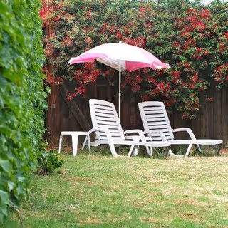 Two white lounge chairs with a pink and white umbrella set up on a grassy lawn, surrounded by green foliage and red flowering bushes with a wooden fence in the background.