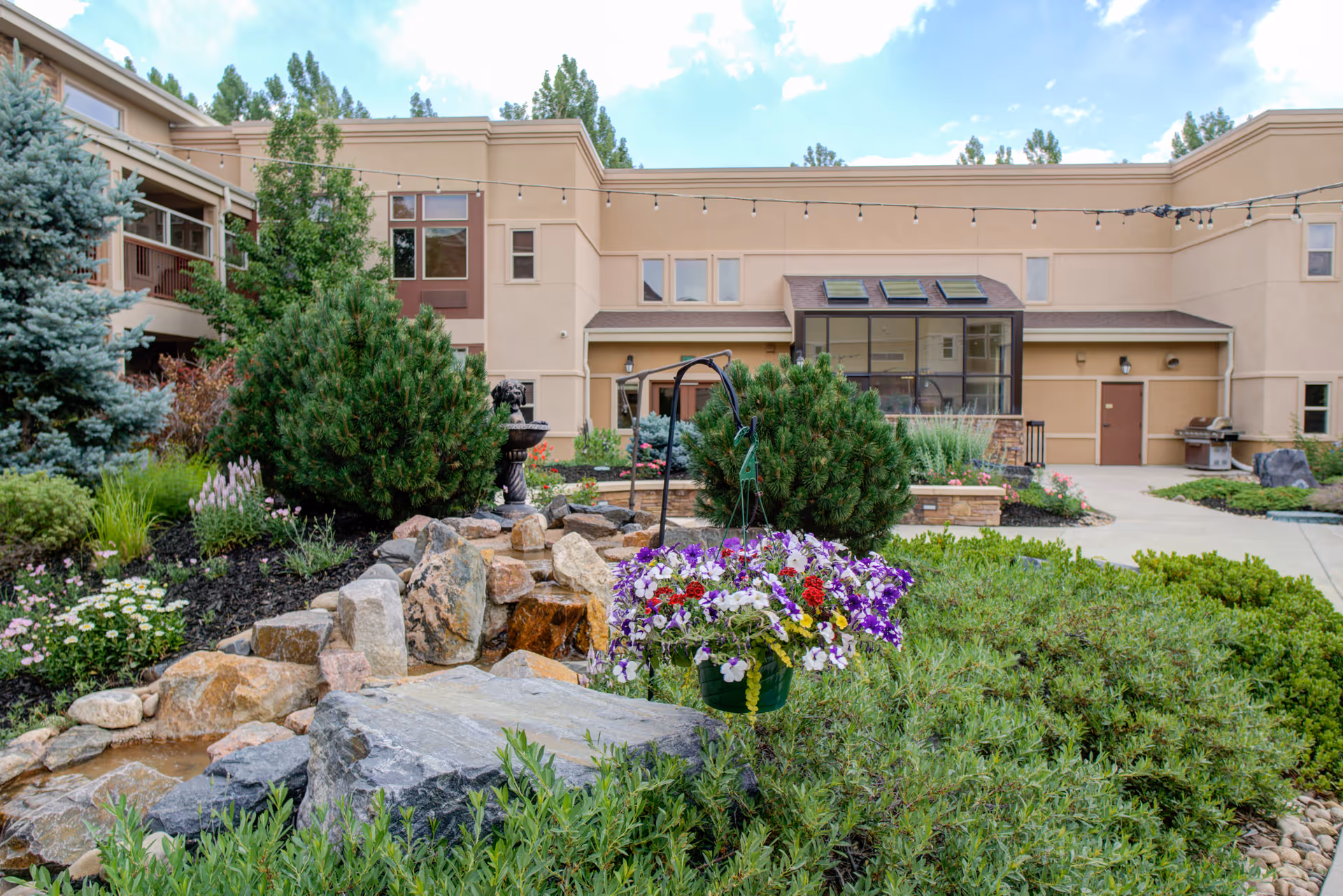 A landscaped garden area in front of a beige senior living facility building with various green shrubs, colorful flowers in a hanging basket, rocks, and a small water feature. The building has multiple windows and a patio area with string lights overhead under a partly cloudy sky.