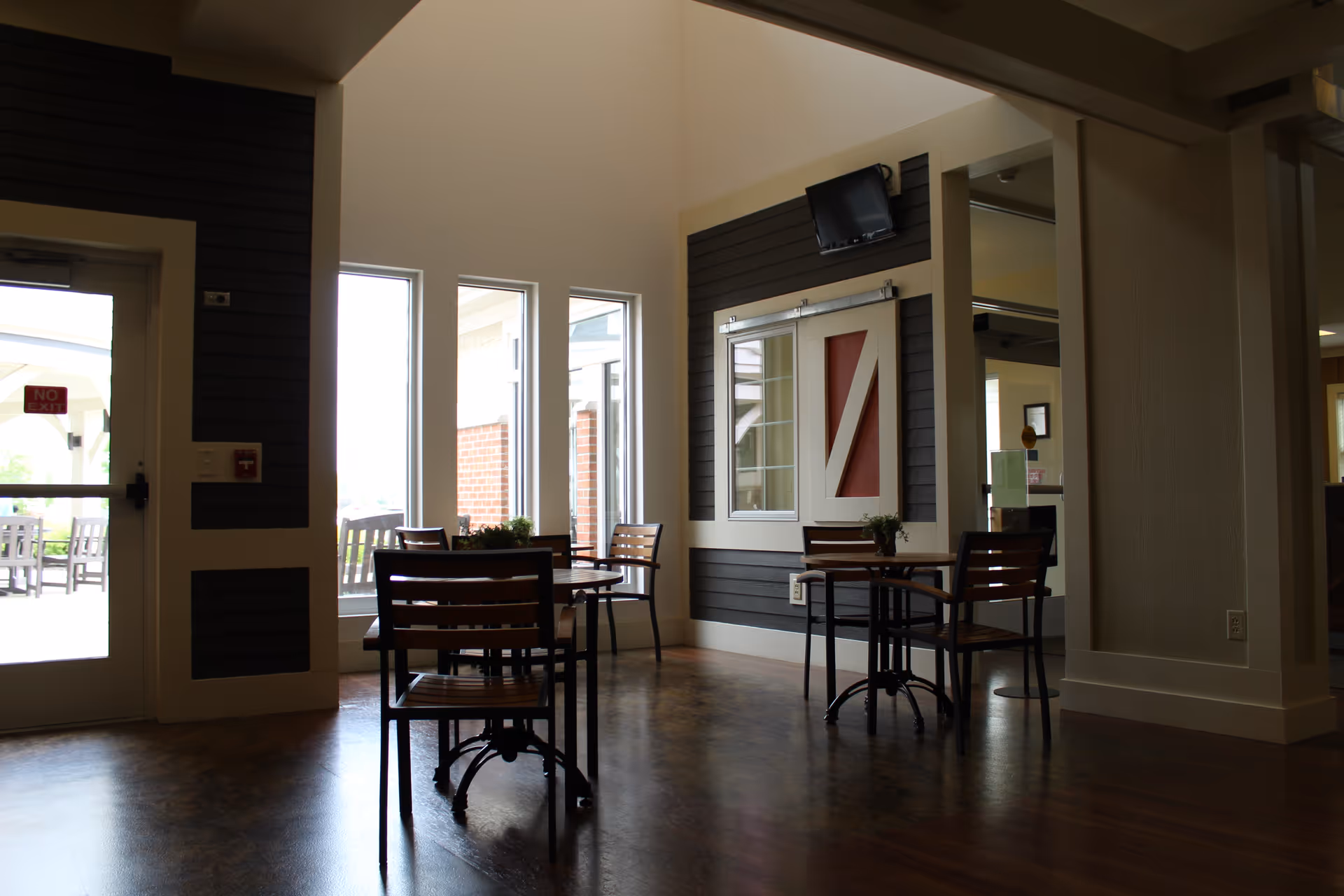 Sunlit common seating area with small round tables and wooden chairs near tall windows and an exterior door.