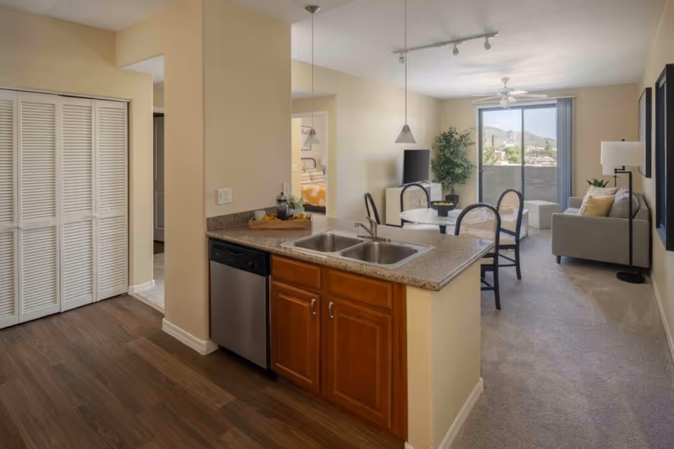 Open-plan apartment interior with a kitchen island and sink in the foreground and a living and dining area with a sofa and balcony in the background.