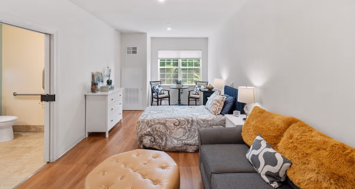 A bright and cozy bedroom with a bed covered in patterned bedding, a gray sofa with orange and patterned pillows, a round beige ottoman, a white dresser with decorative items, and a small table with two chairs near a window. To the left, an open door reveals a bathroom with a toilet and grab bar.