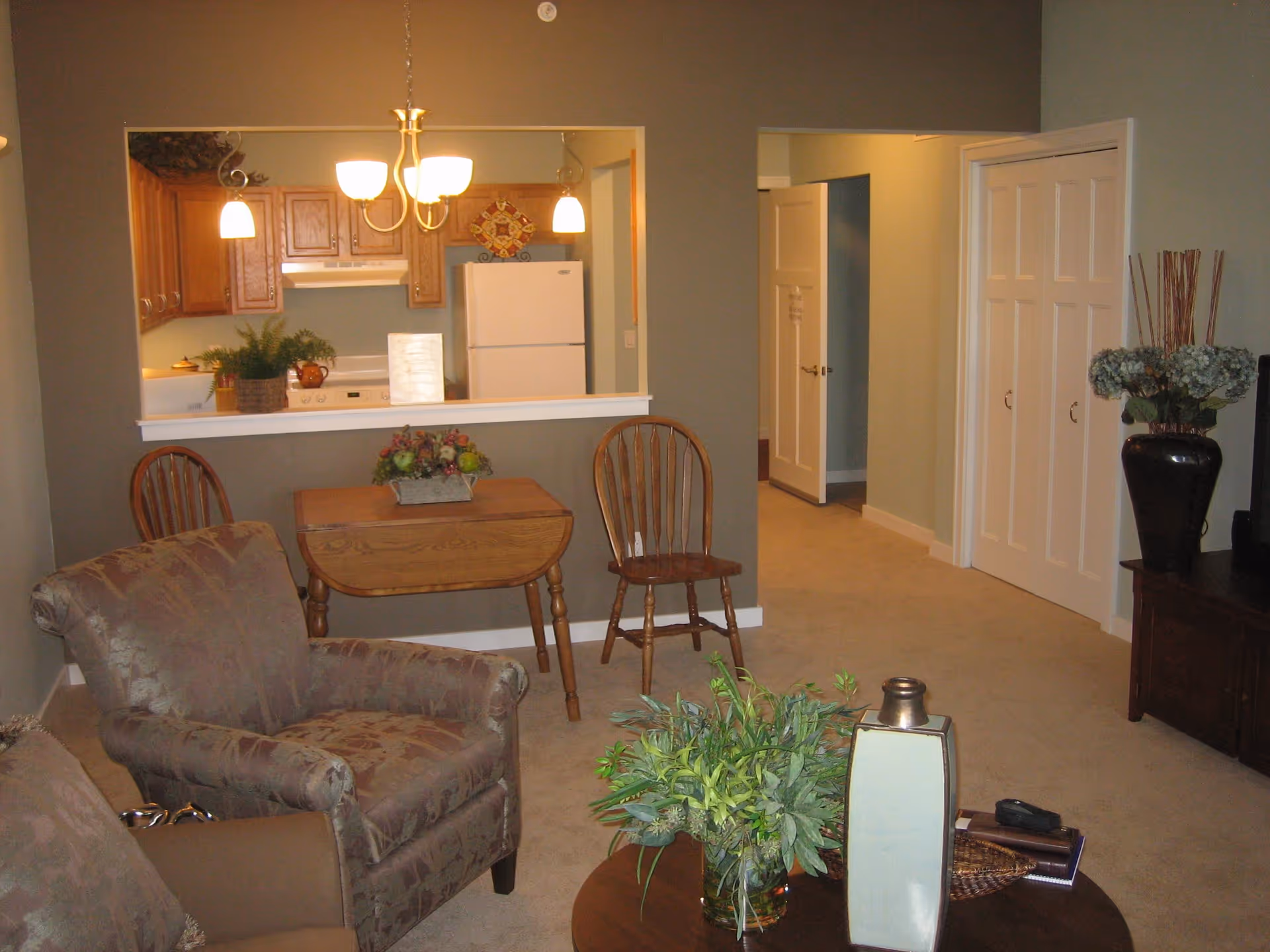 A cozy living room area with two upholstered armchairs and a wooden coffee table adorned with a green plant and decorative items. In the background, there is a small wooden table with two wooden chairs against a wall with a pass-through window opening into a kitchen. The kitchen has wooden cabinets, a white refrigerator, and a stove. To the right, there is a hallway with white doors and a large vase with flowers on a wooden cabinet.