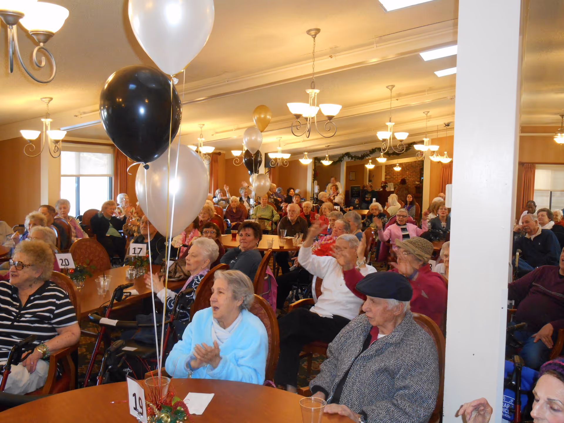 A large group of elderly people seated at round tables in a decorated dining room or common area, some clapping and smiling. There are black, white, and gold balloons tied to the tables, and the room is well-lit with chandeliers and natural light from windows.