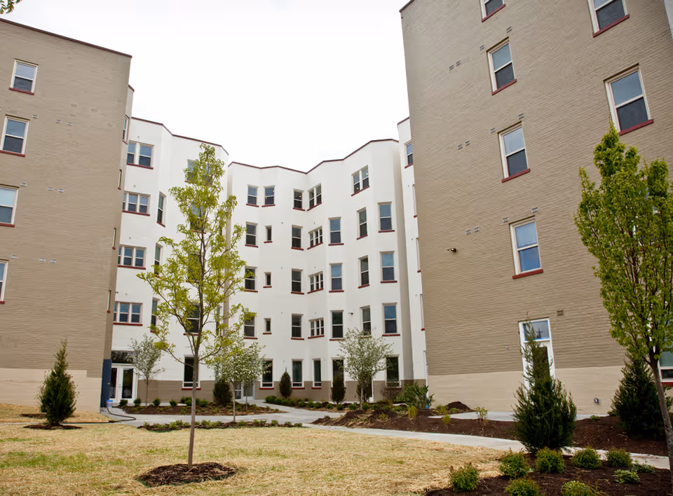 Outdoor courtyard area of The Elberon facility showing a landscaped space with young trees and shrubs surrounded by multi-story beige and white buildings with multiple windows.