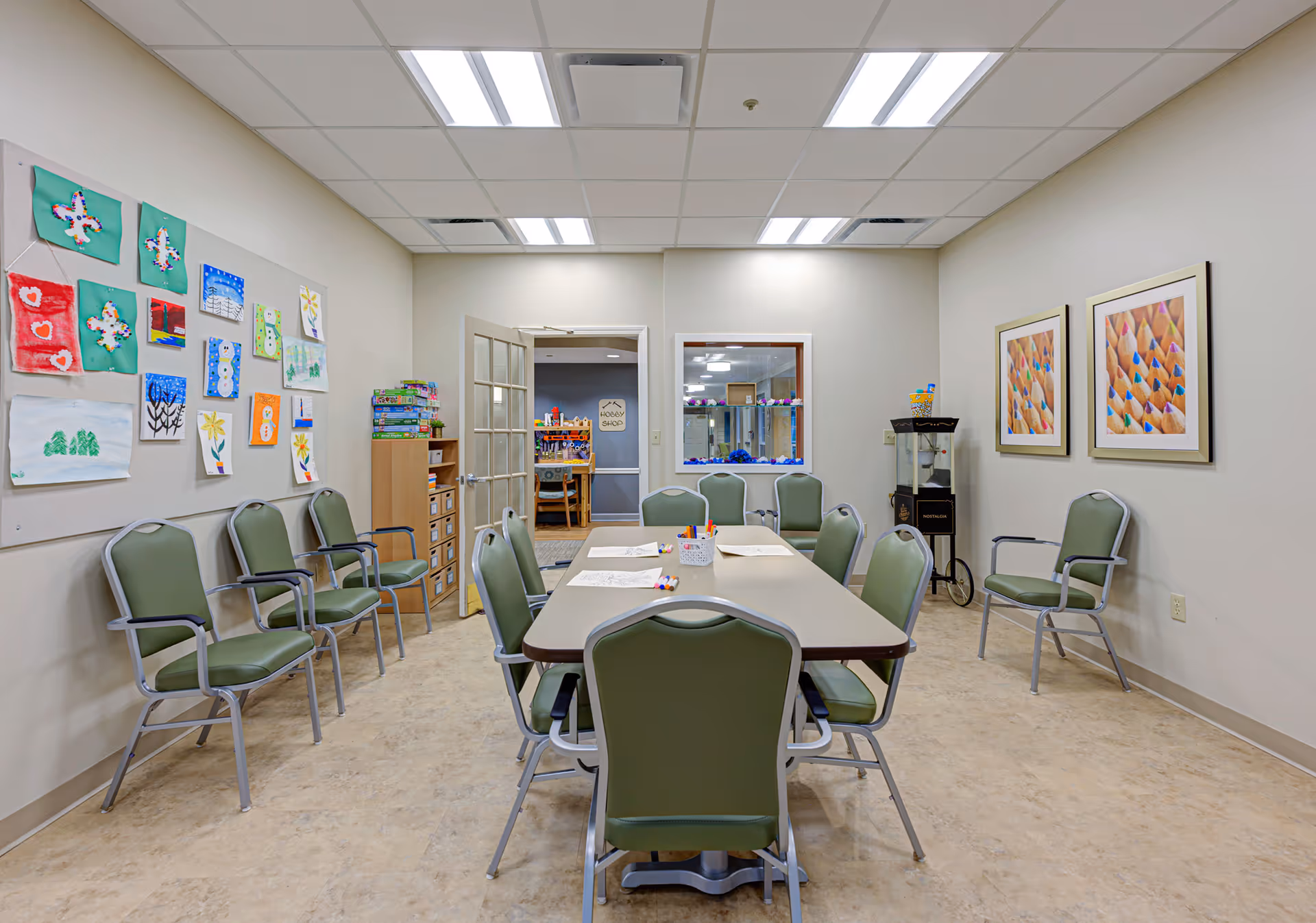 A well-lit activity room with a rectangular table surrounded by green chairs. The walls display colorful children's artwork and framed pictures of colored pencils. There are shelves with board games and a popcorn machine in the corner. A door and a window open to an adjacent room with more games and activities.