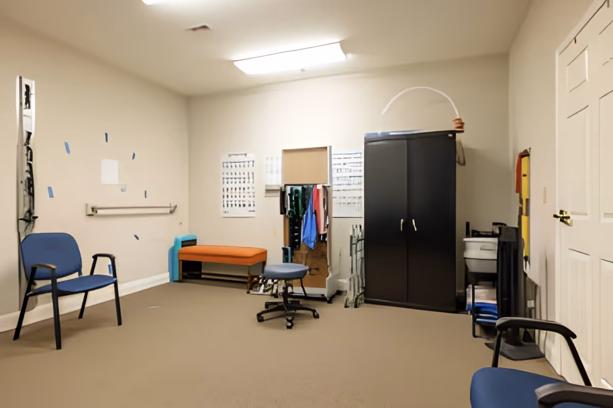 A small room with beige walls and carpeted floor, containing two blue chairs, an orange bench, a black cabinet, a rolling stool, and various items including clothes hanging on a rack and some posters on the wall.