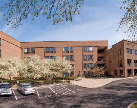 Brick senior living building with parking lot, trees, and blue sky in front.