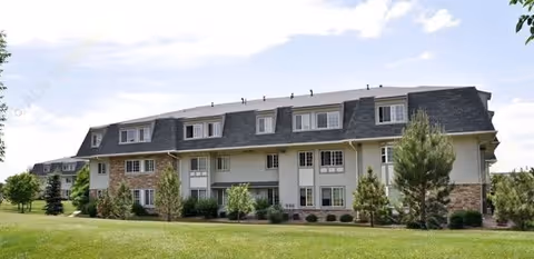 Exterior view of a multi-story senior living facility building named Pinewood Lodge, featuring a combination of stone and siding on the facade, multiple windows, and surrounded by green grass and trees under a partly cloudy sky.