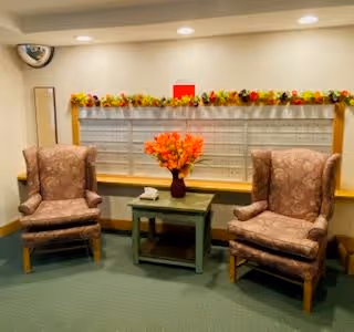 A cozy interior seating area with two upholstered armchairs on either side of a small wooden table holding a vase of orange flowers. The background features a window with closed blinds and a decorative garland above it. The floor is carpeted in green.