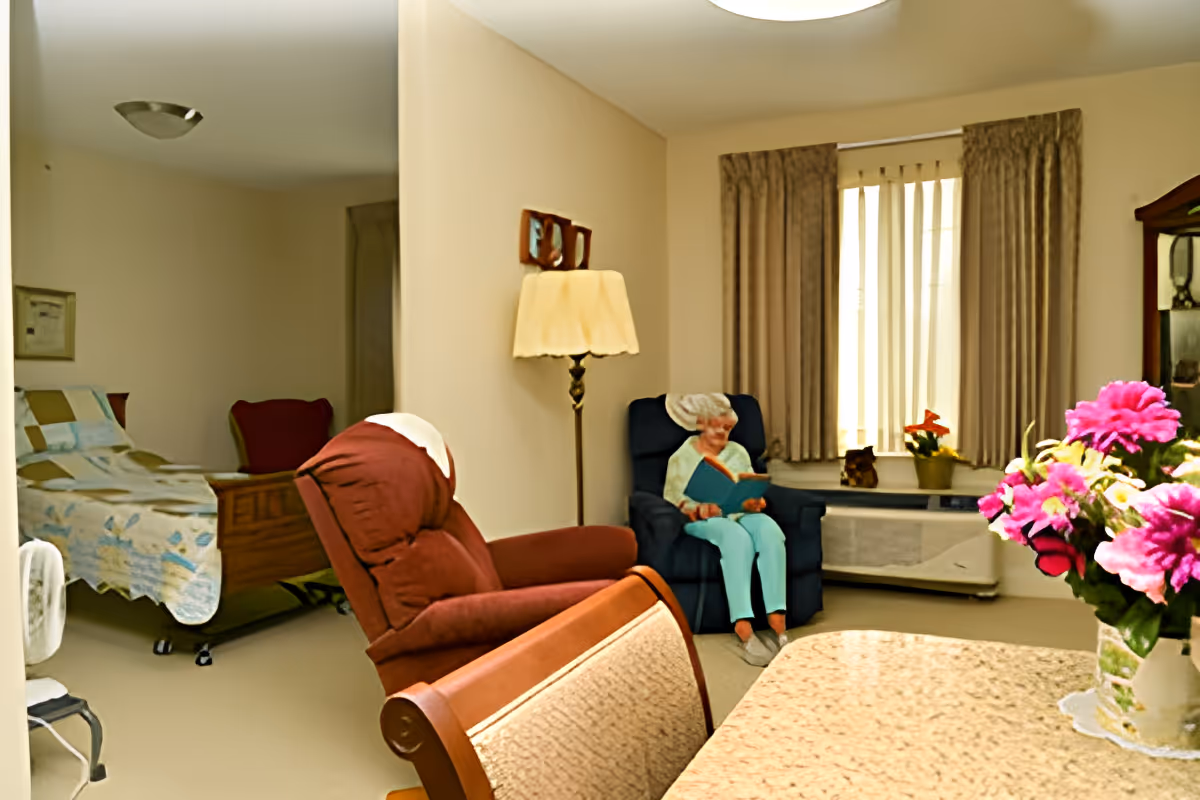 A cozy room in a senior living facility with a bed covered by a patchwork quilt in the background, a red recliner chair in the foreground, and an elderly woman sitting in a blue armchair reading a book near a window with beige curtains. A table with a vase of pink flowers is also visible.