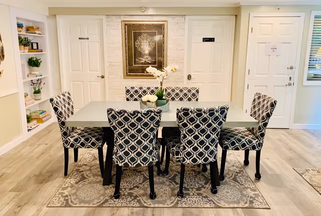 Bright dining room with a rectangular table and six patterned chairs on a rug, decorative wall art and doors in the background.