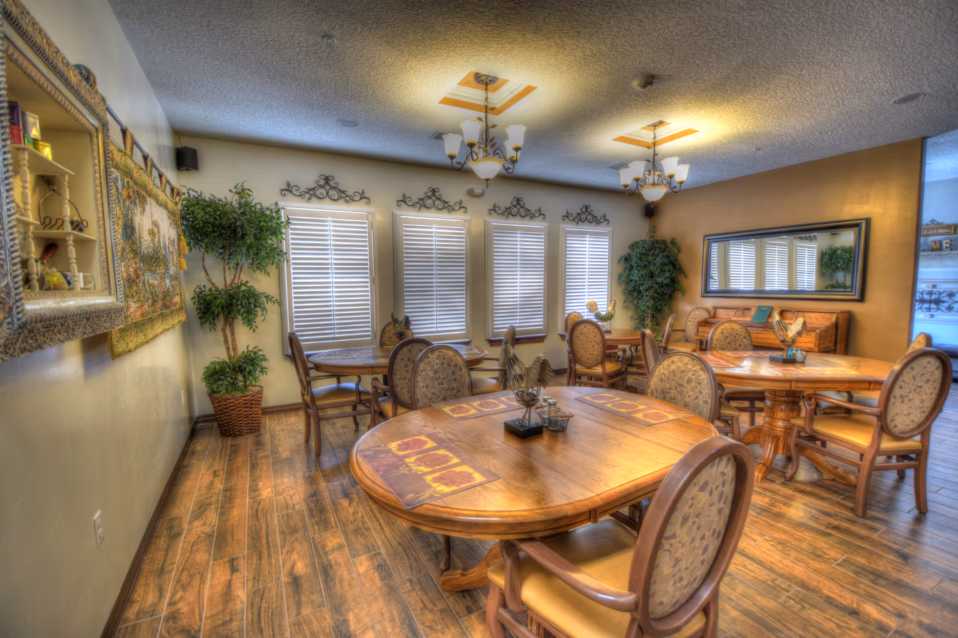 A cozy dining room with several round wooden tables and cushioned chairs arranged neatly. The room features wooden flooring, decorative wall art, potted plants, and windows with white blinds. Two ceiling light fixtures with multiple bulbs provide warm lighting, and a large mirror hangs on one wall.