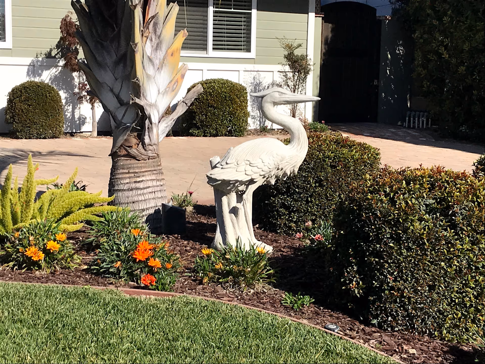 A stone heron statue stands in a landscaped front yard with flowers, shrubs, a palm tree, and a light-green building facade behind it.