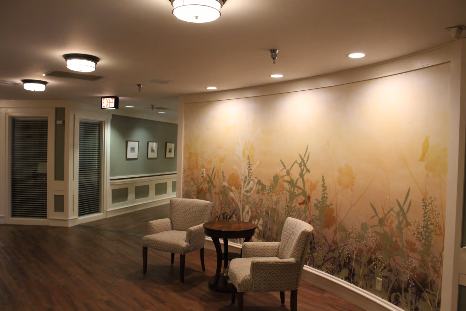 A quiet indoor seating area in a senior living facility with two upholstered armchairs and a small round wooden table between them. The wall behind the chairs features a large mural with a soft floral design in warm tones. The floor is wood, and the ceiling has several recessed lights and round light fixtures. A hallway with framed pictures and an exit sign is visible in the background.