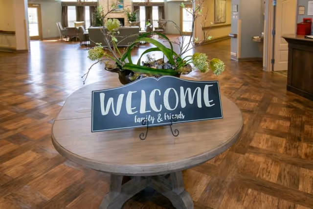 Round wooden table in a facility lobby holding a 'WELCOME family & friends' sign and a floral centerpiece on wood-patterned flooring.