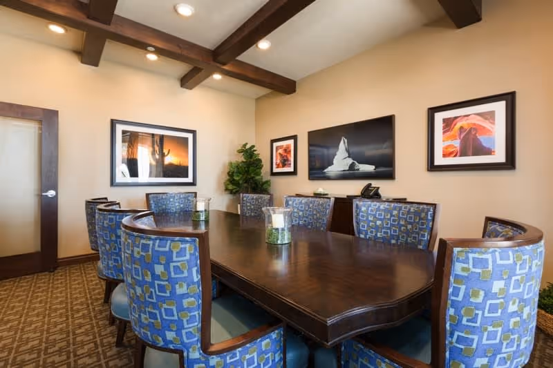 A conference or meeting room with a large dark wooden table surrounded by blue patterned chairs. The room has beige walls with three framed pictures of natural landscapes and a green potted plant in the corner. The ceiling features exposed wooden beams and recessed lighting.