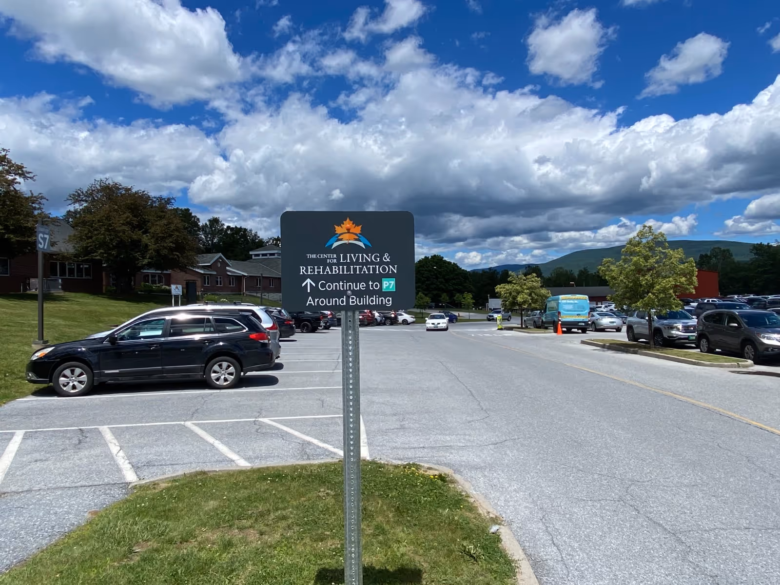 Parking lot and driveway of The Center for Living & Rehabilitation showing a directional sign, parked cars, and buildings under a partly cloudy sky.